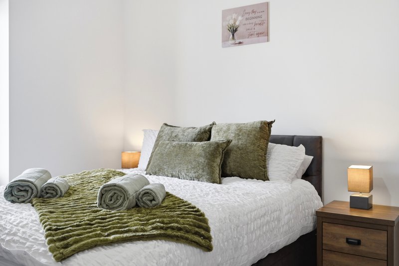 Bedroom with double bed, olive green bedding, rolled towels, and wooden nightstand with lamp