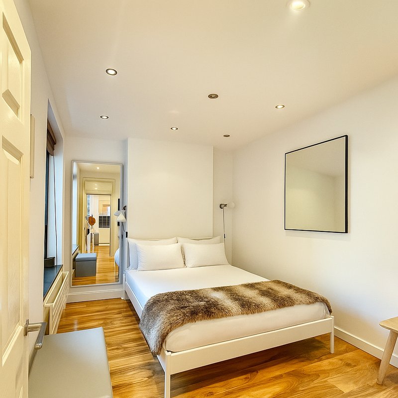 Bright bedroom with white bed, wooden floor, recessed lighting and framed mirror on wall