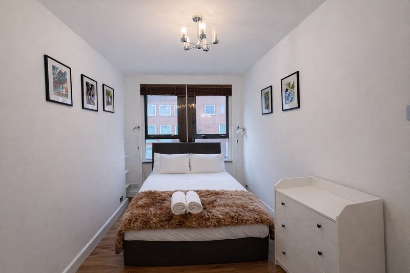 Narrow bedroom with double bed, brown throw, framed wall art, window and white dresser