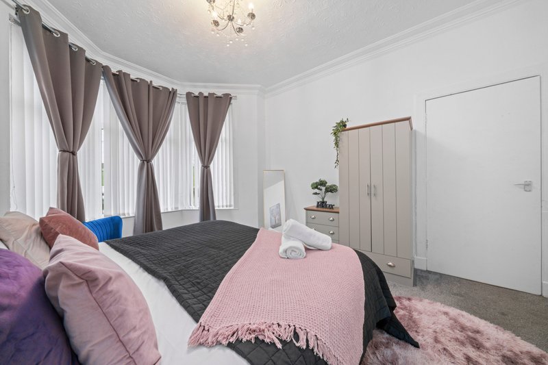 Bright bedroom with double bed, colorful cushions, pink throw blanket, and floor-to-ceiling windows with brown curtains