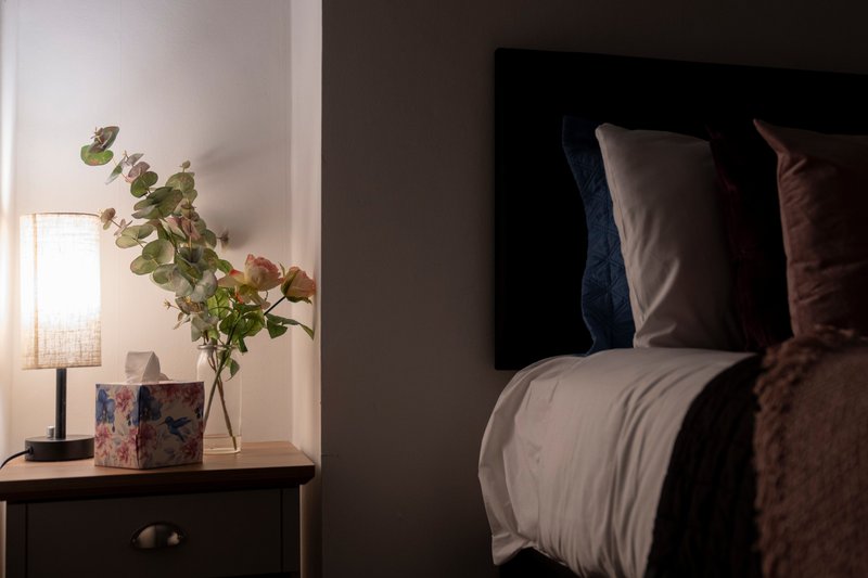 Bedroom corner with bedside table, lamp, vase with greenery, and made bed with pillows