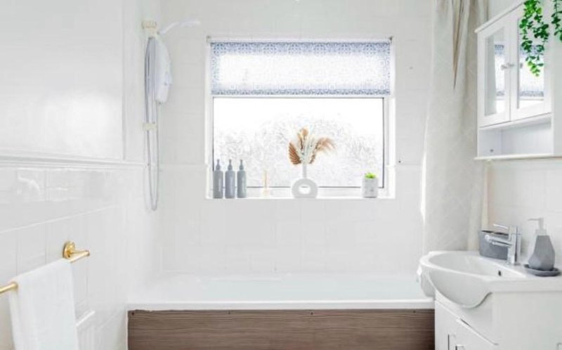 Bright modern bathroom with white walls, wooden bathtub panel, sink and window with natural light