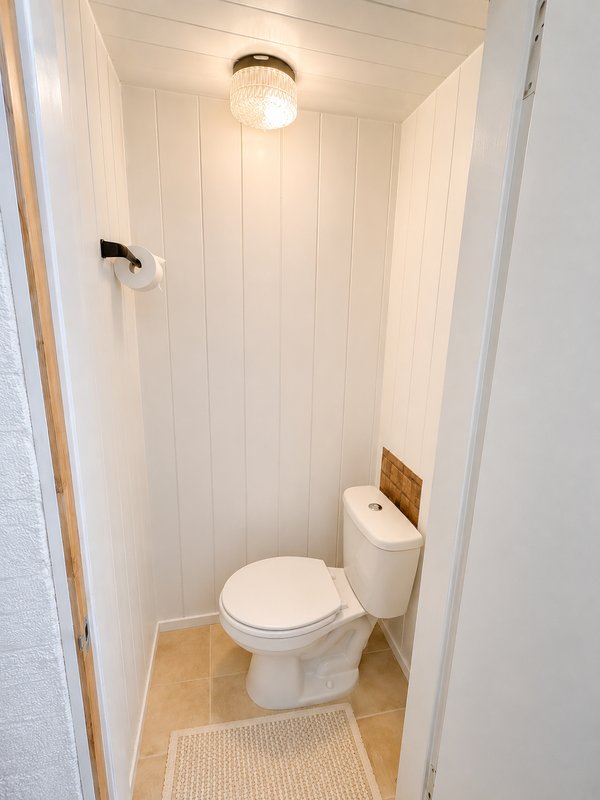 Small bathroom with toilet, white panelled walls, ceiling light and beige floor mat