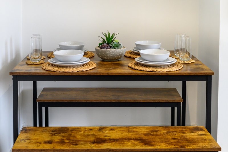 Wooden dining table set with place settings, woven placemats, potted plant, and glassware