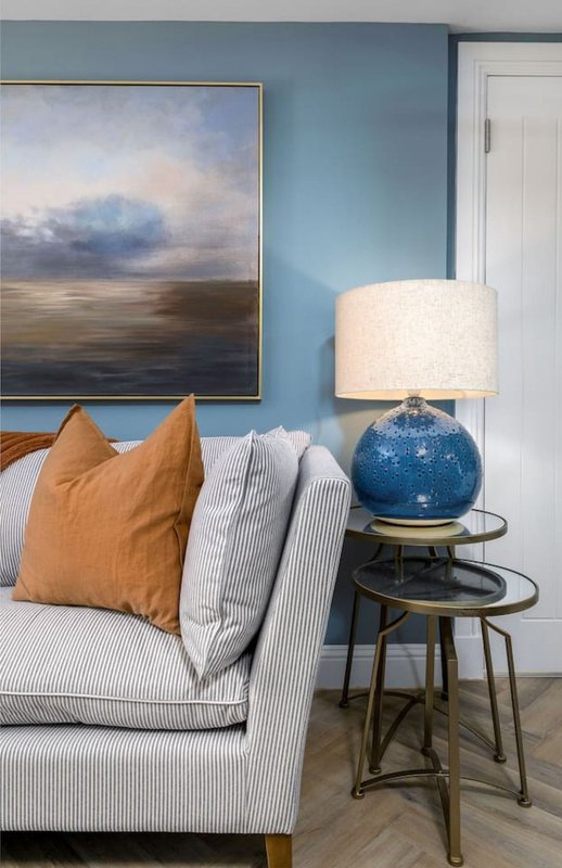 Living room with light blue walls, striped sofa, orange cushion, landscape artwork and blue table lamp