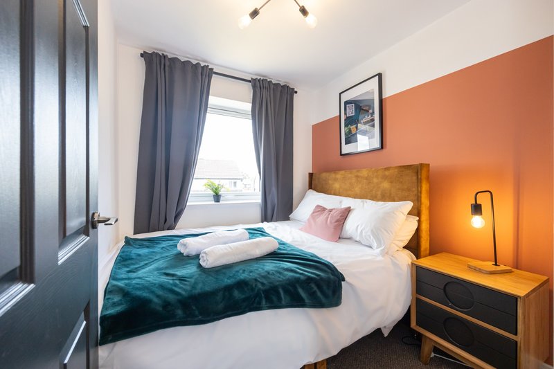 Bedroom with double bed, teal bedding, wooden headboard, orange accent wall, and window with curtains