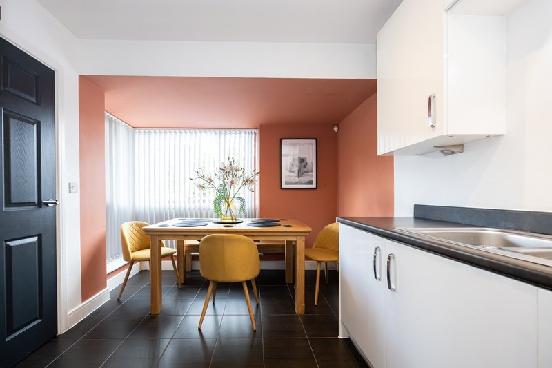 Modern kitchen and dining area with wooden table, yellow chairs, orange accent wall and window.