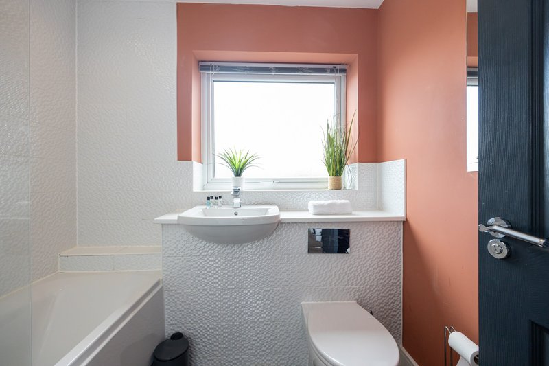 Modern bathroom with coral accent wall, white sink, toilet, and window with plants
