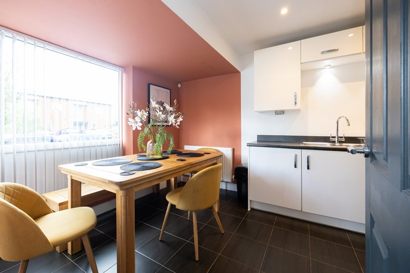 Modern kitchen and dining area with wooden table, yellow chairs, white cabinetry and pink accent wall
