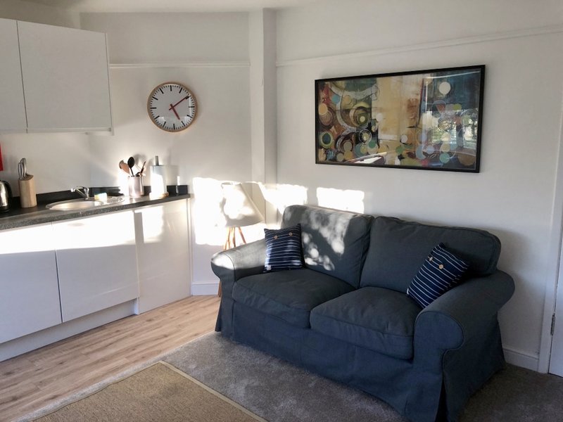 Open-plan living space with white kitchen, black sofa, wall clock and framed artwork