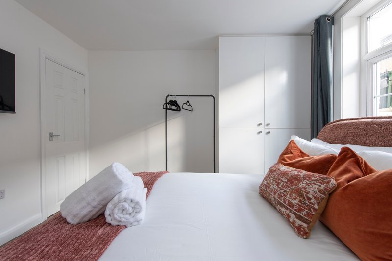 Modern bedroom with white bed, orange and burgundy bedding, wardrobe and window with curtains