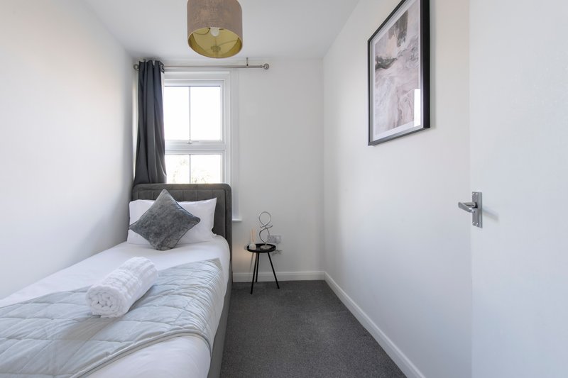 Compact bedroom with single bed, grey cushion, window with curtains, and framed wall art