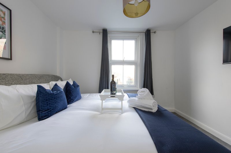 Modern bedroom with double bed, navy cushions, window with grey curtains, and bedside table with wine bottle