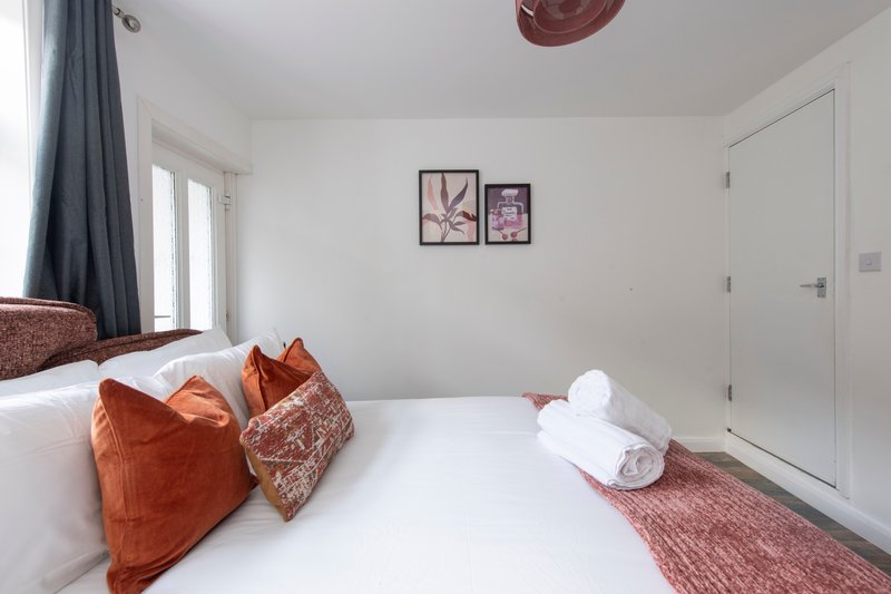 Bright bedroom with white bed, orange and patterned cushions, rolled towels, and framed wall art