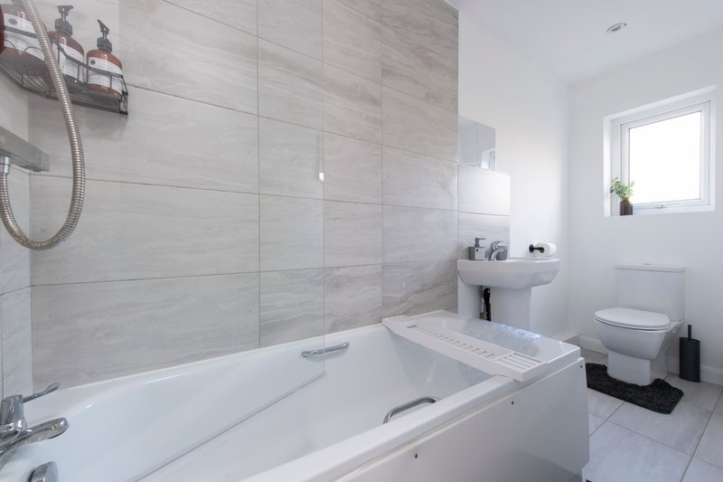 Modern bathroom with white bathtub, grey tile walls, pedestal sink and toilet