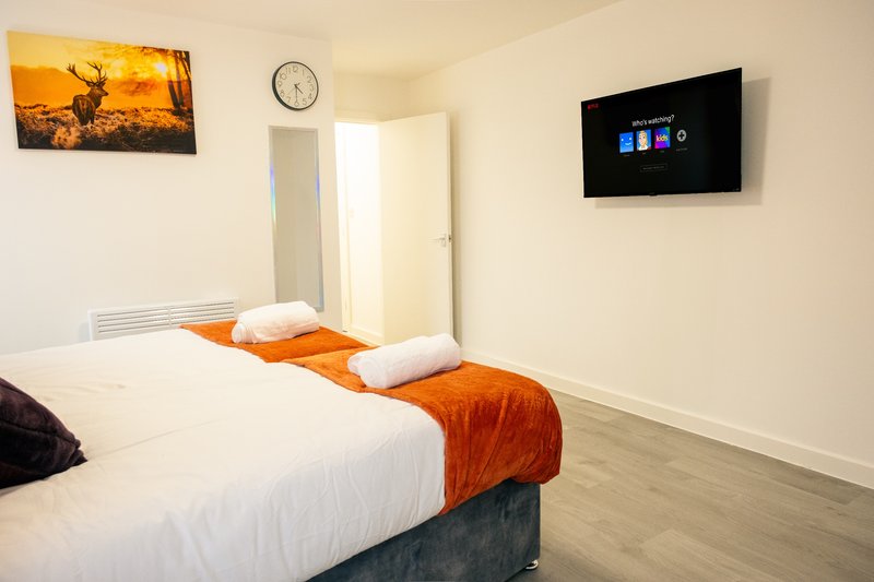Bedroom with double bed, white pillows, orange bedding, wall-mounted TV and clock