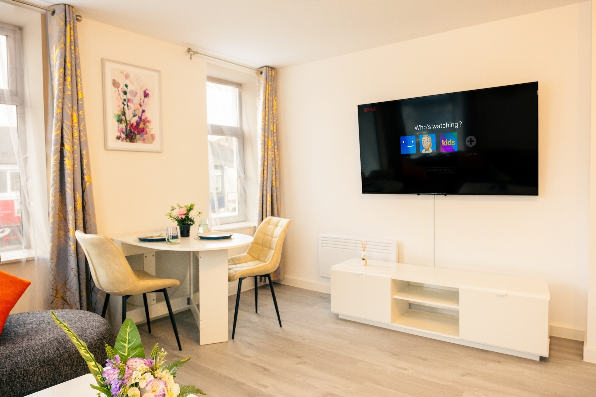 Bright living room with wall-mounted TV, dining table, chairs, and windows with yellow curtains