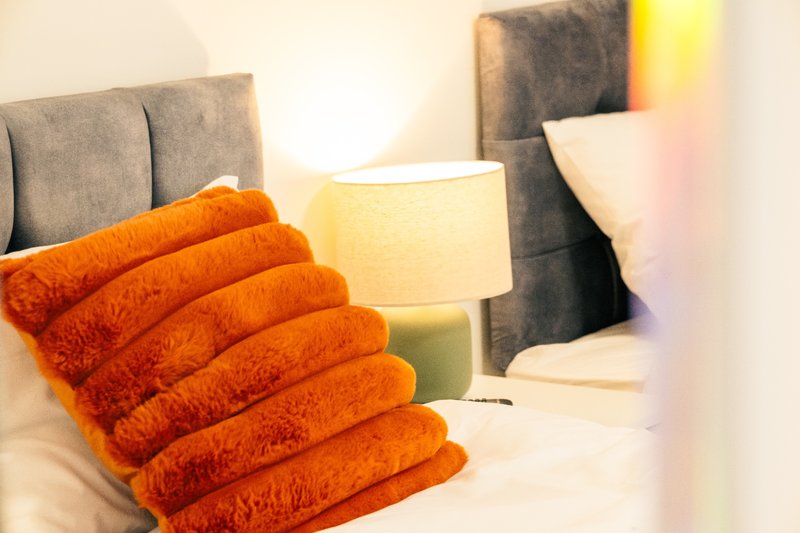 Bedroom with grey upholstered headboard, orange towels on bed, table lamp and pillows