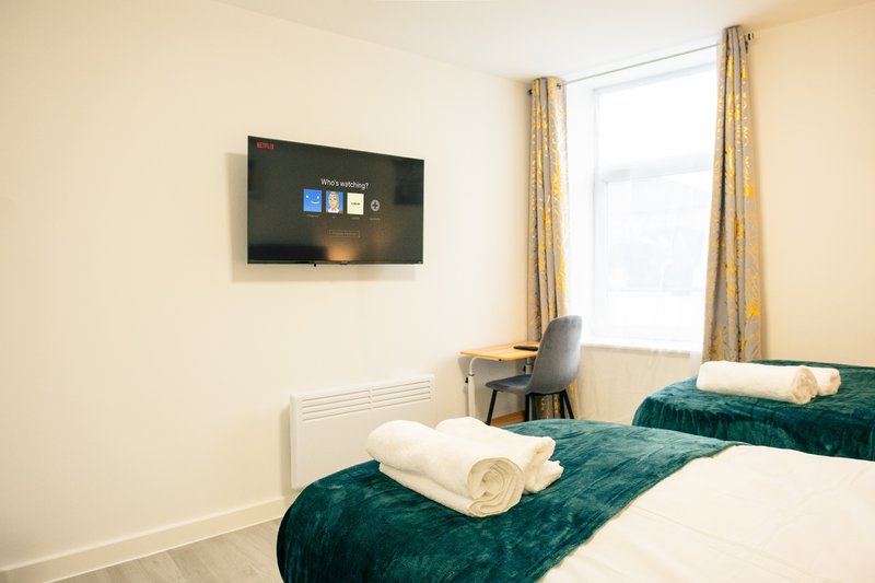 Bedroom with two beds, teal bedding, rolled towels, wall-mounted TV, and window with gold curtains