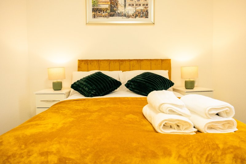 Bedroom with mustard-yellow headboard, bed, black pillows, white rolled towels, and framed wall art