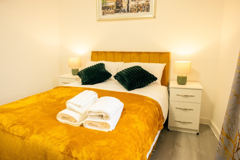Bedroom with yellow bedspread, mustard headboard, green pillows, white folded towels, and bedside tables