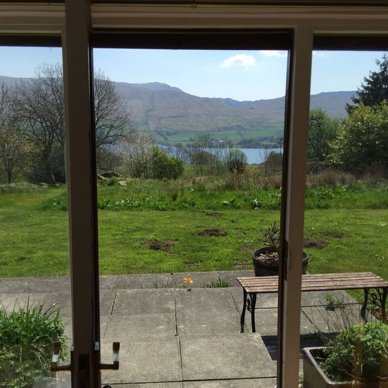 View through glass doors to patio, garden, and lake with mountains in background