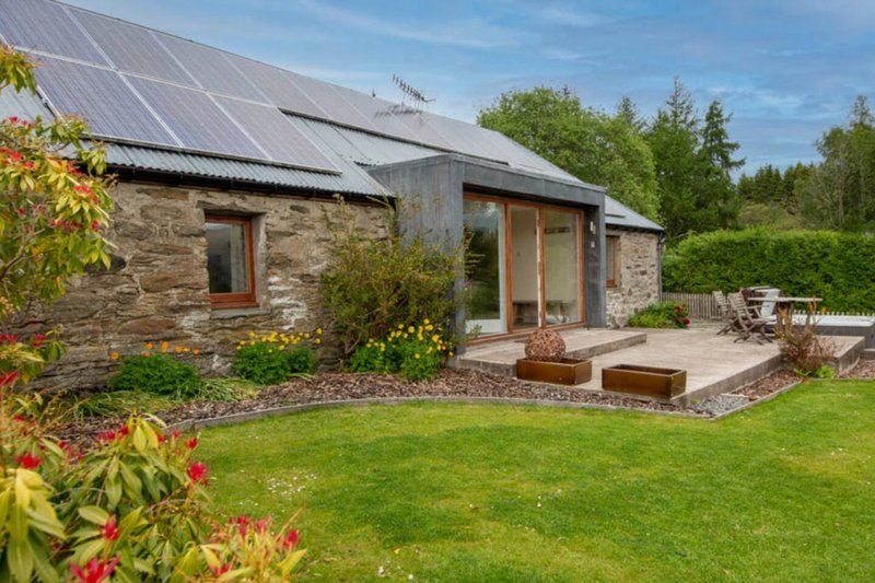 Modern stone cottage with solar panels, wooden-frame windows, patio seating and manicured garden
