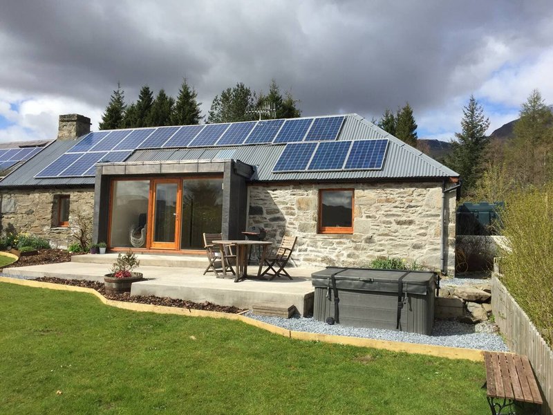 Modern stone cottage with solar panels, orange entrance doors, patio seating and garden