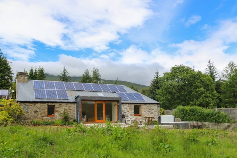 Modern stone cottage with solar panels, wooden entrance, surrounded by green fields and trees