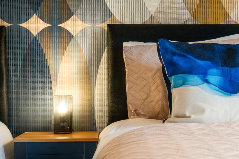 Modern bedroom with patterned wallpaper, wooden nightstand, bedside lamp and blue pillows