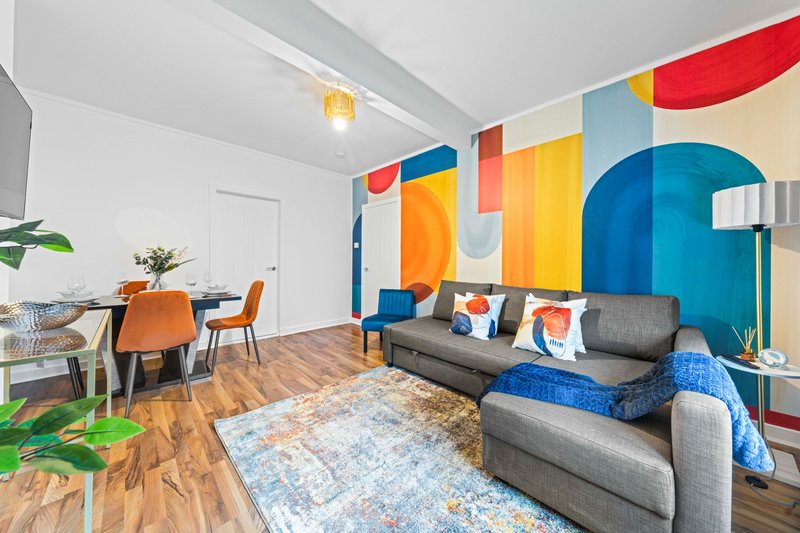 Open-plan living room with colorful geometric wall mural, gray sofa, dining area with orange chairs, wooden flooring