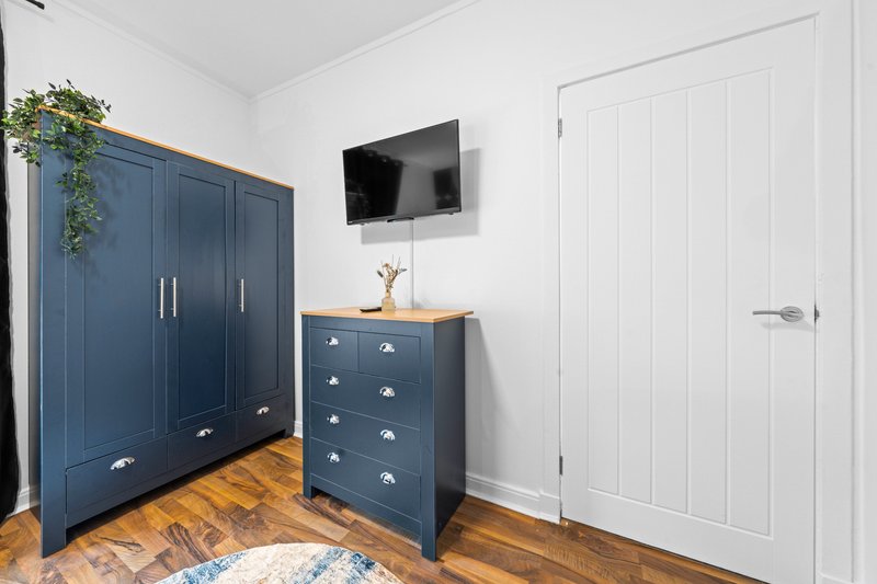Bedroom with blue wardrobe, dark chest of drawers, wall-mounted TV, wooden floor and white door