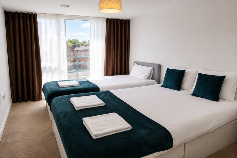 Bright bedroom with two double beds, teal bench seating, dark curtains and large windows overlooking street