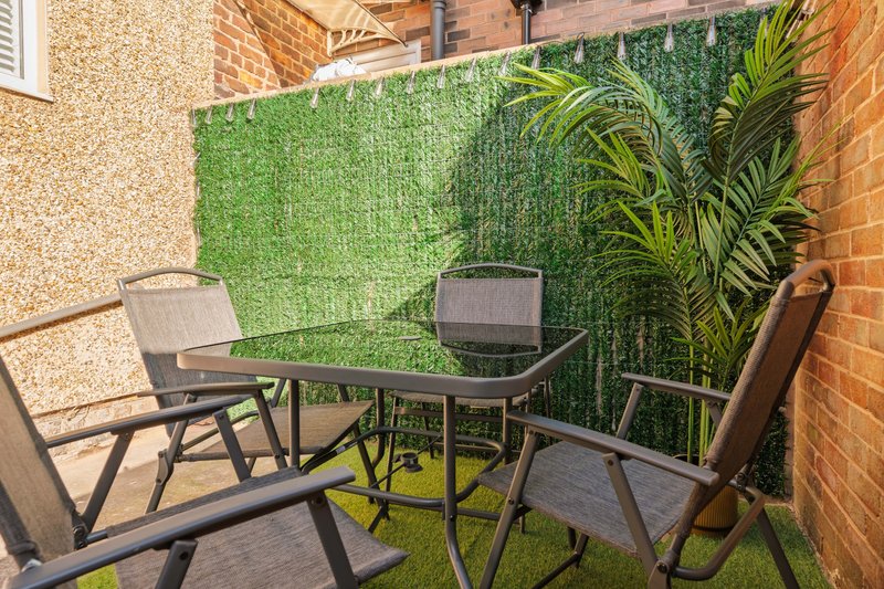Small brick patio with artificial turf, metal outdoor table, grey chairs and green plant screening