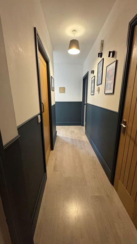 Narrow hallway with two-tone wall paneling, wooden floor, ceiling light fixture and framed wall art