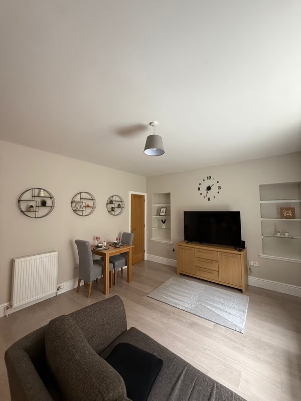 Open-plan living room with grey armchair, wooden TV unit, dining area and wall clock