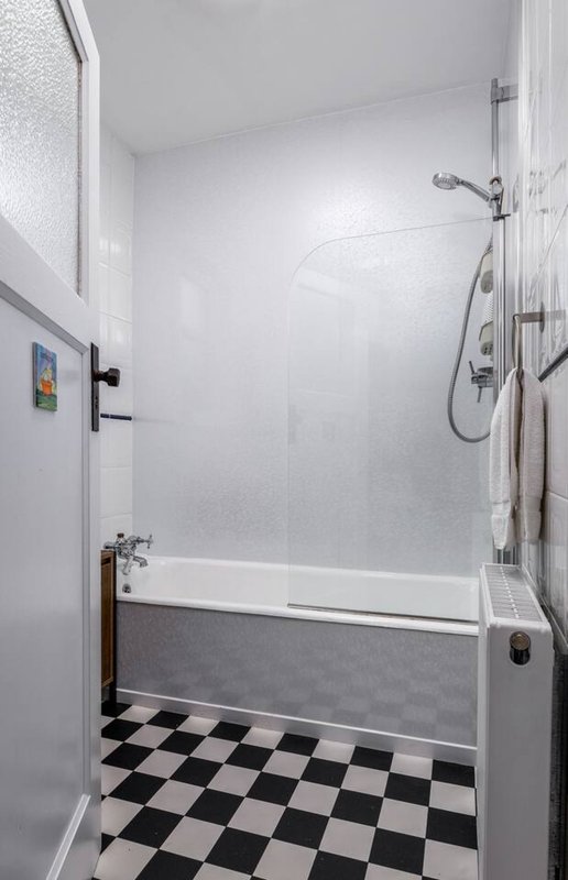 Clean bathroom with white tile walls, black and white checkered floor, bathtub with shower enclosure and handheld showerhead