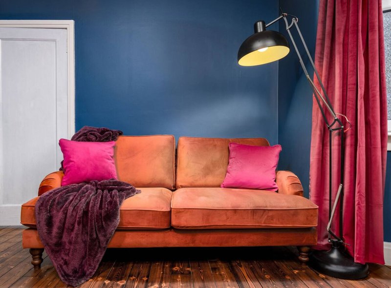 Living room with orange sofa, pink cushions, burgundy throw blanket, black floor lamp and blue walls
