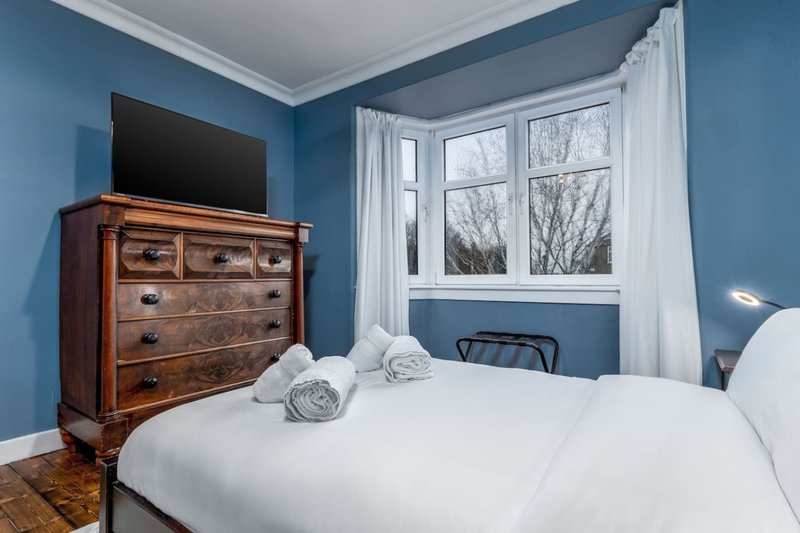 Bright bedroom with blue walls, white bedding, wooden dresser, TV, and large window with curtains