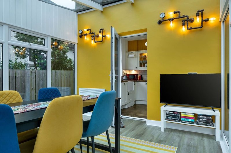 Open-plan living and dining area with yellow walls, blue and yellow chairs, TV, and garden view
