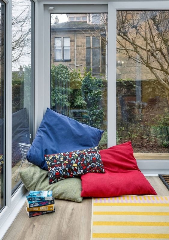 Window seat with blue and red cushions, striped throw, stacked books and view of garden
