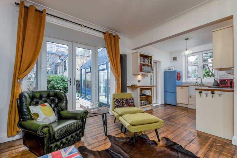 Open-plan living area with green leather armchair, yellow ottoman, kitchen, and large windows with orange curtains