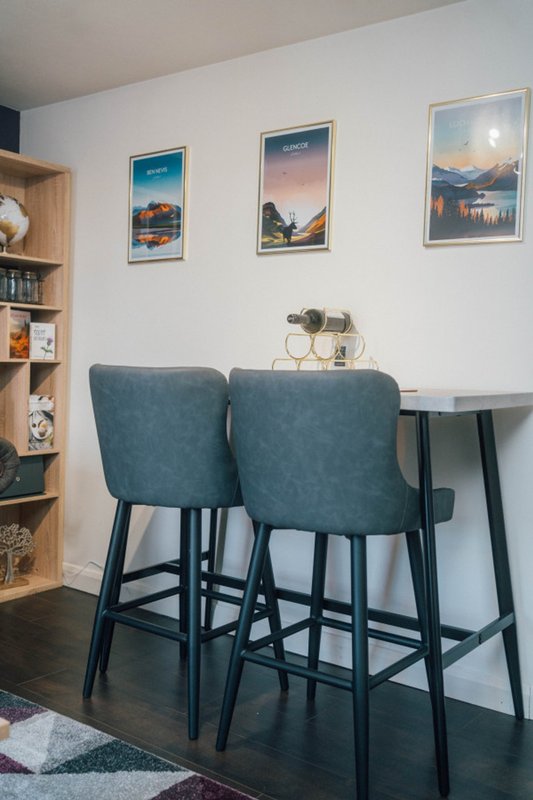 Modern dining area with two teal upholstered bar stools, small black table, and framed landscape artwork on white walls