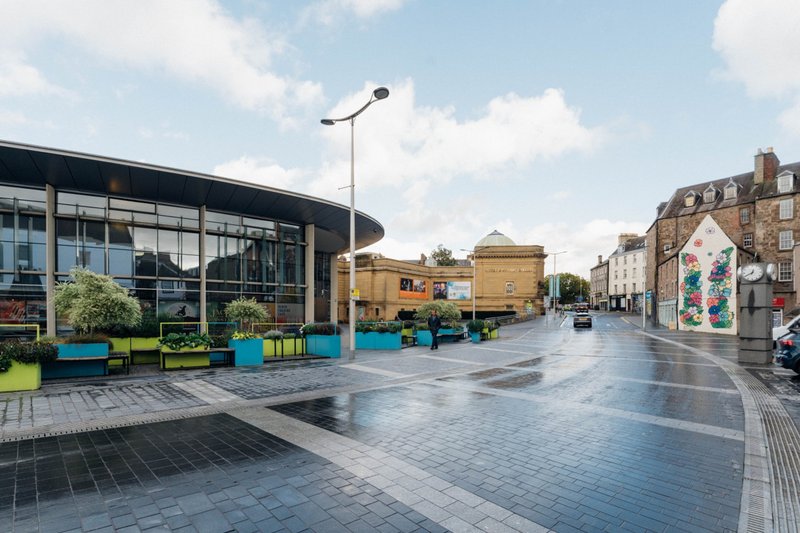 Modern building exterior with large windows, street view, paved plaza, and surrounding architecture