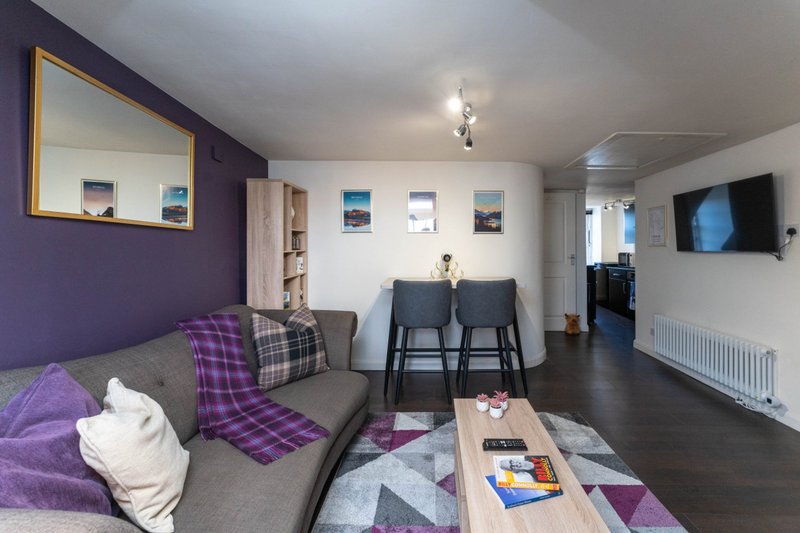 Open-plan living room with gray sofa, purple accents, dining table, and wall-mounted TV