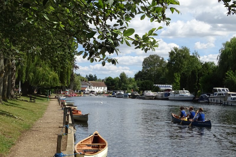 Riverside scene with moored boats, tree-lined bank, and waterfront buildings across the water