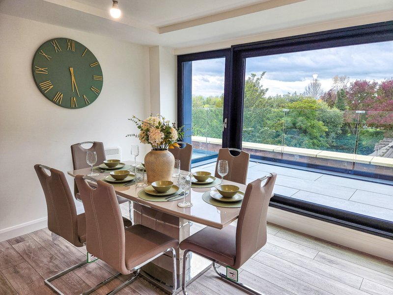 Modern dining room with glass table, upholstered chairs, wall clock and large windows overlooking landscape