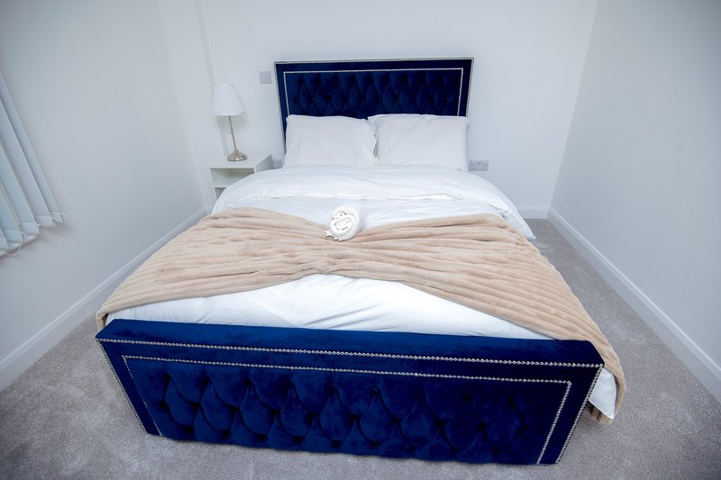 Bedroom with blue upholstered bed, white bedding, beige throw blanket and bedside lamp