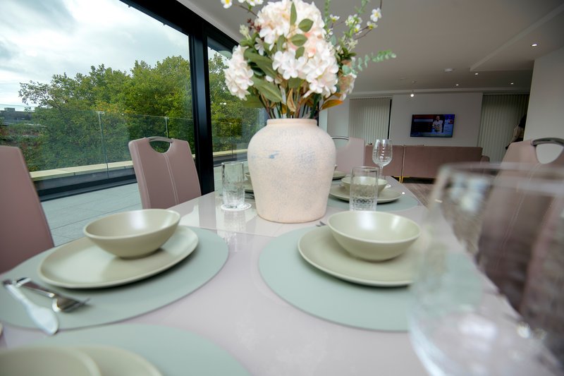 Modern dining table with place settings, white vase with flowers, and countryside view through large windows