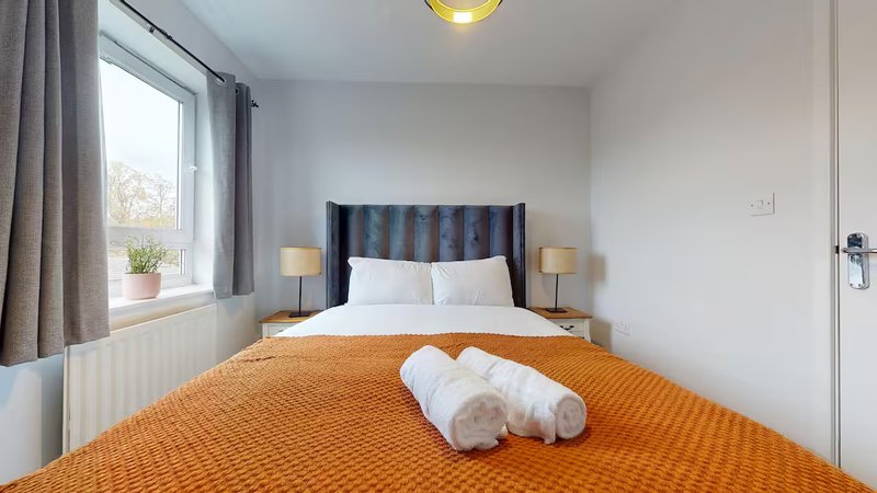 Bright bedroom with double bed, orange bedspread, striped headboard, and window with curtains